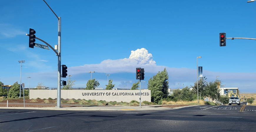 Depicted is the UC Merced campus in the foreground, with smoke from a distant wildfire seen behind it on a blue sky.
